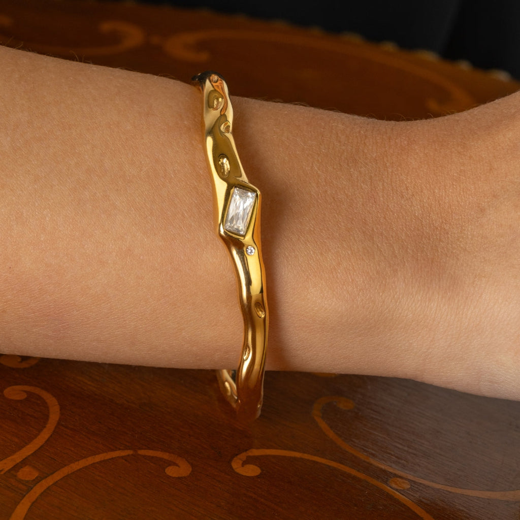 Gold bracelet with a rectangular gemstone on a wrist against a patterned background