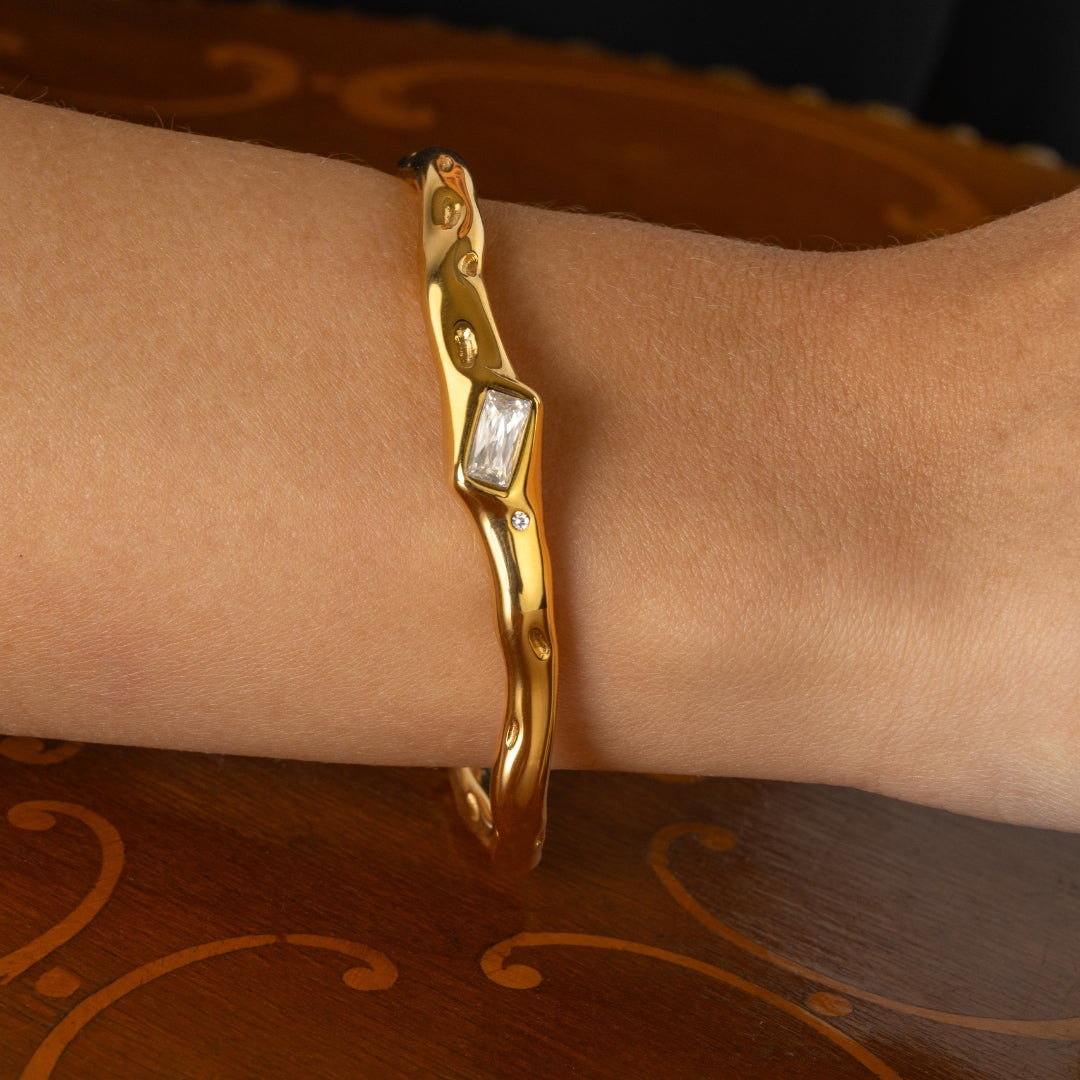 Gold bracelet with a rectangular gemstone on a wrist against a patterned background
